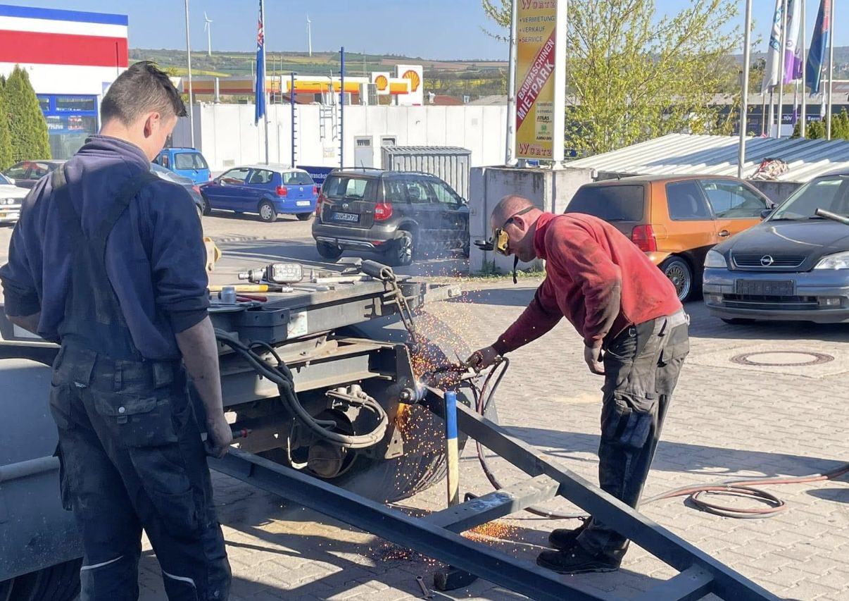 Detailansicht des Motorraums eines Lastwagens, der sich offenbar in Wartung oder Reparatur befindet. Man sieht einen großen, beigen Lüfter, Teile des Kühlers und die blauen Scheinwerfer des Fahrzeugs. Der Motorraum ist offen und gibt den Blick auf technische Komponenten frei.