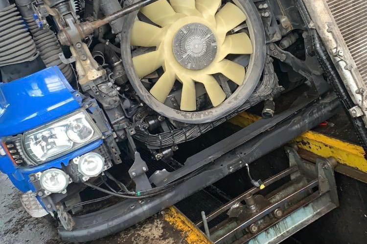 Detailansicht des Motorraums eines Lastwagens, der sich offenbar in Wartung oder Reparatur befindet. Man sieht einen großen, beigen Lüfter, Teile des Kühlers und die blauen Scheinwerfer des Fahrzeugs. Der Motorraum ist offen und gibt den Blick auf technische Komponenten frei.